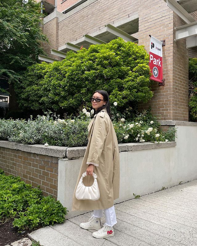 Jovem posando em frente a muro com plantas, ela está de lado usando óculos escuros pretos e com expressão séria. Ela usa blusa bege e calça jeans clara.