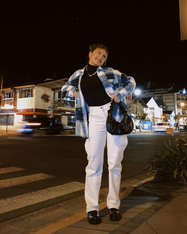 Jovem posando na rua com as mãos na cintura, usando camisa xadrez com calça branca e bolsa preta. Sua expressão é sorridente.