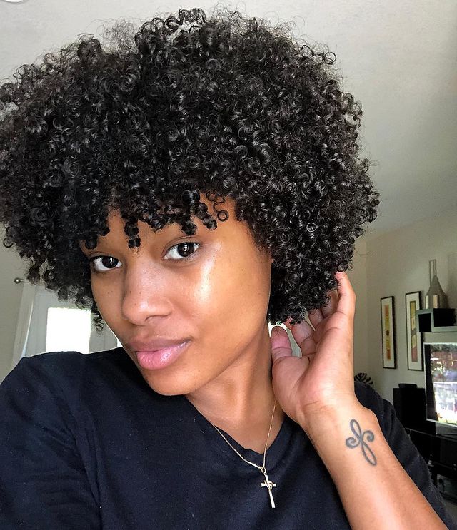 cabelo big chop Selfie de uma mulher. Ela usa uma camiseta preta, colar com pingente de cruz e cabelo solto. Ela está com a mão esquerda no cabelo, com o rosto levemente de perfil, olha para a câmera mas não sorri.