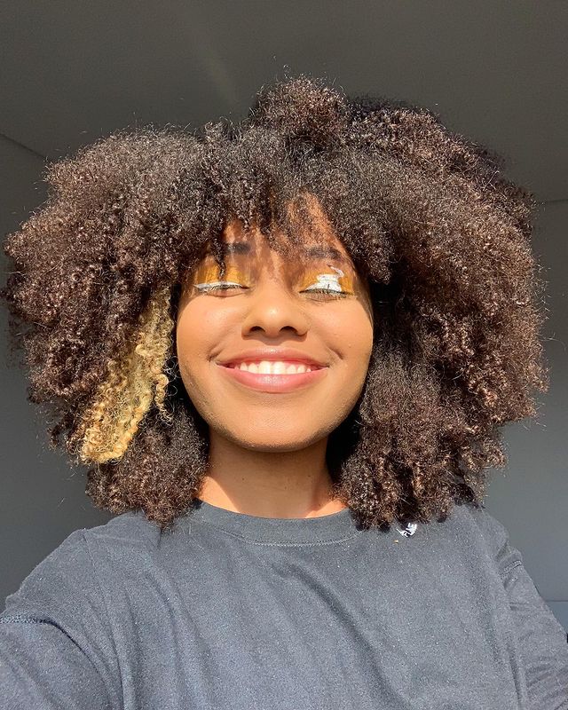 cabelo crespo em camadas Selfie de uma mulher sorrindo. Ela usa uma camiseta preta e cabelo crespo solto.