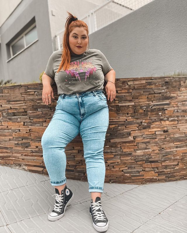 calca-jeans-skinny Foto de uma mulher encostada em um muro. Ela usa uma camiseta cinza, calça jeans skinny e All Star preto de cano alto. Ela olha para a câmera e sorri levemente.