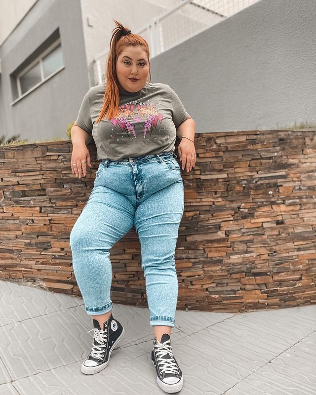 Foto de uma mulher encostada em um muro. Ela usa uma camiseta cinza, calça jeans skinny e All Star preto de cano alto. Ela olha para a câmera e sorri levemente.