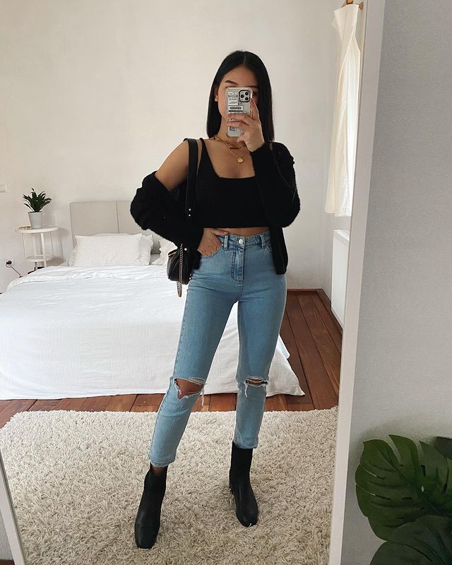 calça-skinny Selfie no espelho de uma mulher. Ela usa um top cropped preto, cardigan preto, calça jeans skinny rasgada nos joelhos, bota preta e bolsa preta.