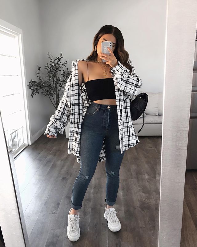 calça skinny look Selfie no espelho de uma mulher mostrando o look. Ela usa uma calça skinny escura, cropped preto, camisa xadrez oversized branco e preta, bolsa preta e tênis branco.