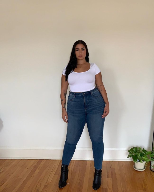 calca-skinny Foto de uma mulher. Ela usa uma camiseta branca, calça skinny azul e bota preta. Ela olha para a câmera e não sorri.