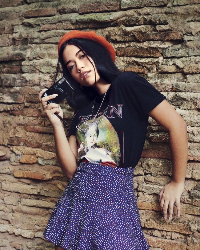 camisa de banda com boina Jovem apoiada na parede posando com camiseta preta, saia azul e boina vermelha.