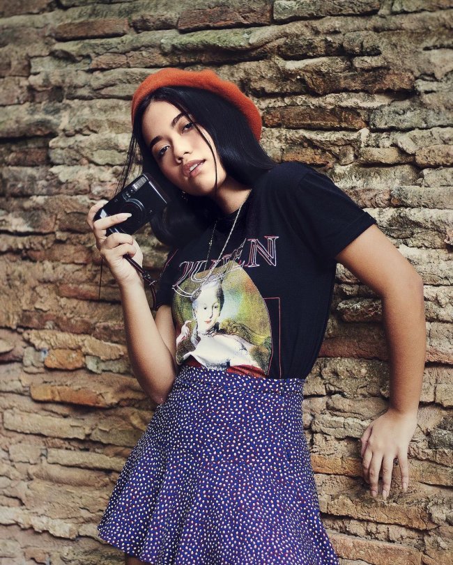 Jovem apoiada na parede posando com camiseta preta, saia azul e boina vermelha.