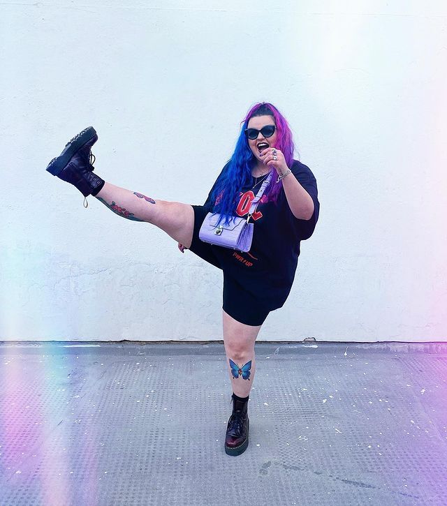 camisa de banda com coturno Jovem fazendo pose com uma perna levantada, com cabelo colorido usando óculos escuro em fundo branco.