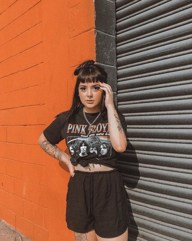 camisa de banda e short Jovem posando em frente parede laranja com uma das mãos no rosto e usando camiseta de banda amarrada na cintura.
