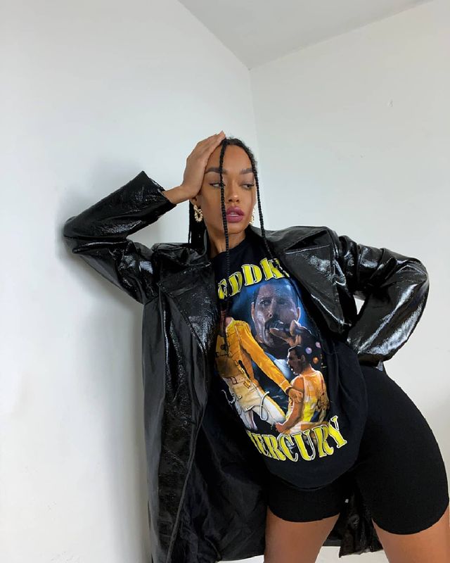 camiseta de banda com sobretudo Jovem apoiada na parede branca com uma das mãos na cabeça, ela usa camiseta de banda e sobretudo brilhante preto.
