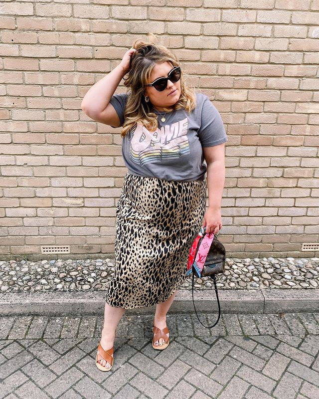 camiseta de banda com animal print Jovem posando na rua em frente a parede de tijolos, uma de suas mãos está no cabelo, ela usa óculos preto.