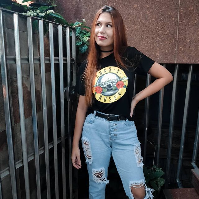 Jovem posando com camiseta de banda, mão na cintura, expressão sorridente calça jeans.