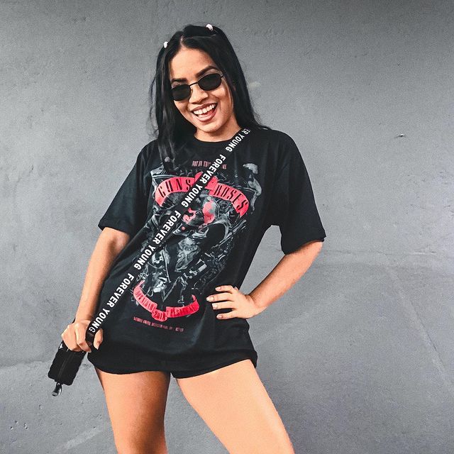 camiseta de banda com short curto Jovem posando com o corpo levemente inclinado para o lado direito e uma das mãos na cintura, ela usa camiseta de banda, short preto e óculos escuros.