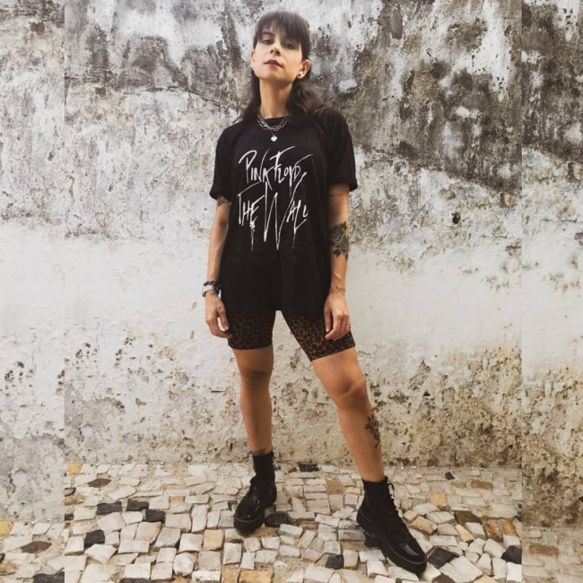 camiseta de banda com short Jovem posando em frente a parede branca com manchas pretas, ela tem uma expressão séria, e usa camiseta de banda com short biker, ambos na cor preta.