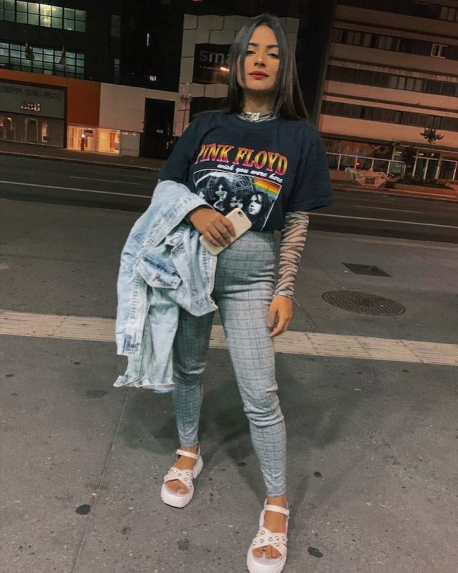 Jovem com calça cinza xadrez posando no meio da rua com camiseta de banda e jaqueta jeans dobrada na mão.