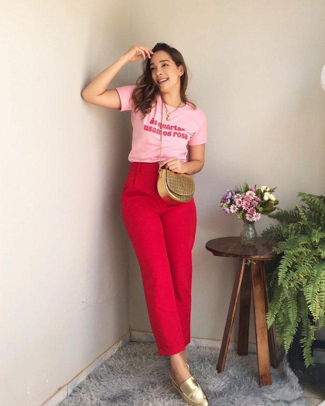 Jovem posando encostada na parede branca usando camiseta rosa com calça vermelha ao lado de plantas verdes e em um banco de madeira marrom.