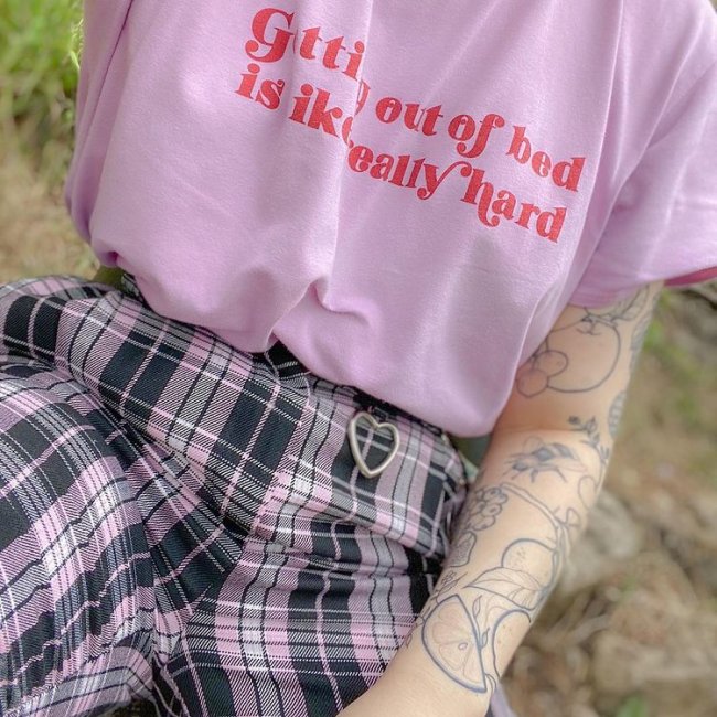 Na imagem só podemos ver a camiseta rosa com uma frase escrita em vermelho. A pessoa também uma calça xadrez lilás e preta.