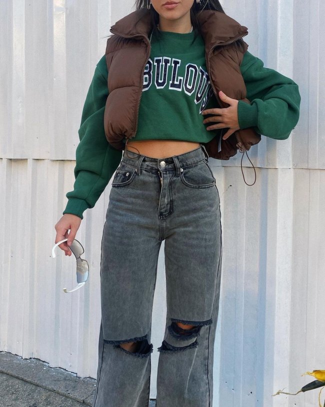 Foto do look de uma garota. Ela usa uma blusa de moletom verde escuro, um colete puffer marrom, calça modelo wide leg cinza. Ela está com a mão esquerda no colete e segura um óculos de sol com a mão direita.