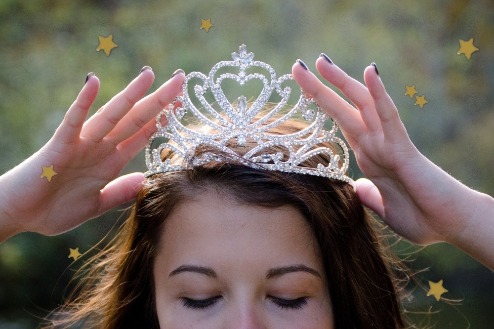 Jovem colocando em si mesma uma tiara de princesa, só conseguimos ver seus olhos fechados enquanto ela faz isso. O restante do rosto não aparece.