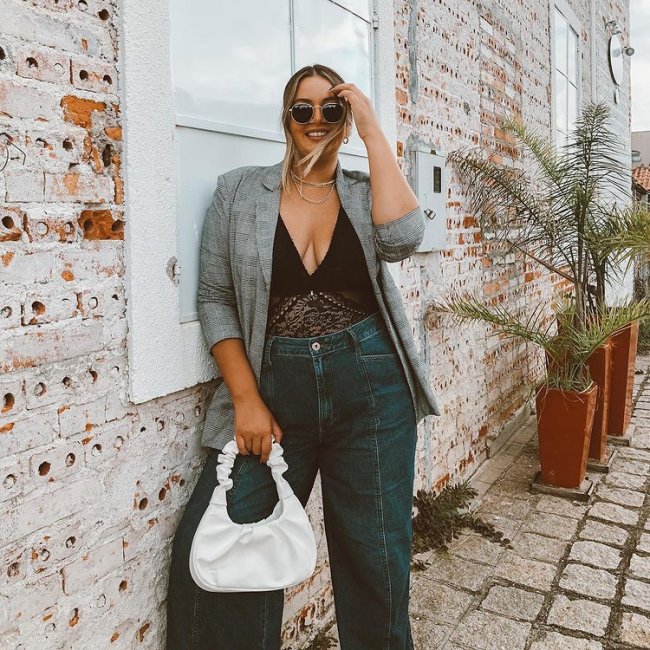 influenciadora com a bolsa jw pei gabbi branca Jovem posando com blazer cinza e body preto, uma de suas mãos está apoiada na cabeça, sua expressão é sorridente e ela está segurando uma bolsa branca.