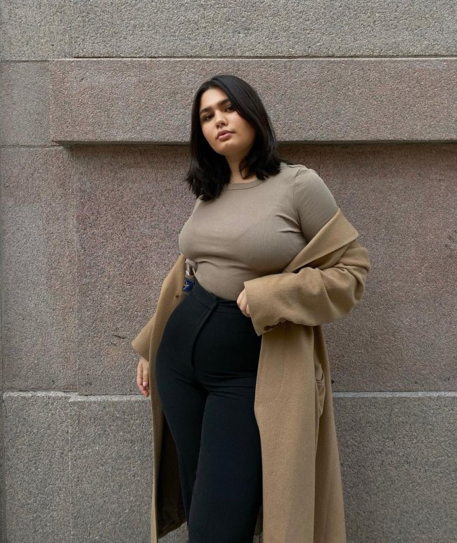 jeans-skinny Foto de uma mulher na rua. Ela usa uma camiseta bege, casaco bege e calça skinny preta. Ela olha para a câmera e não sorri.