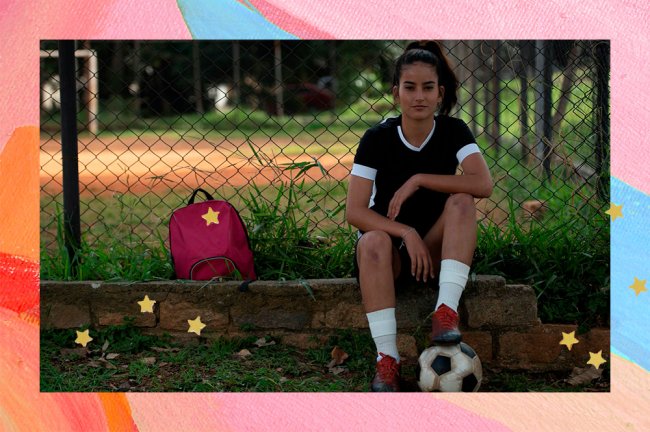 A jogadora de futebol Kezia Sousa está sentada ao lado de um campo, com uma bola embaixo do pé