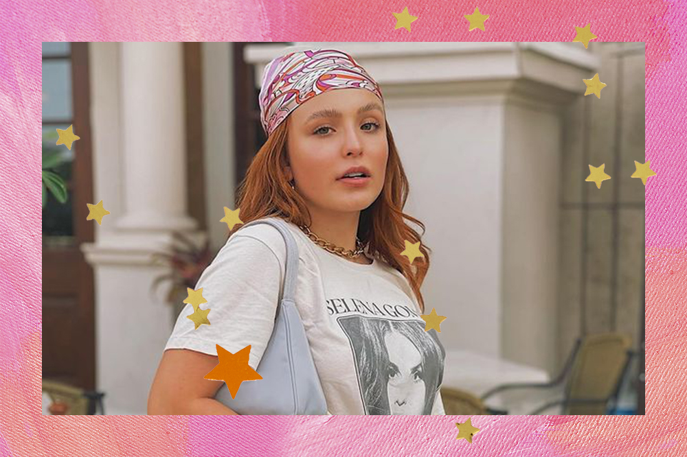 Montagem com foto da Larissa Manoela em fundo rosa com estrelas douradas e laranjas. Ela está usando uma camiseta branca da Selena Gomez, uma bolsa azul pastel pendurada em um dos ombros e um lenço rosa na cabeça. Sua expressão facial é séria em direção à câmera.