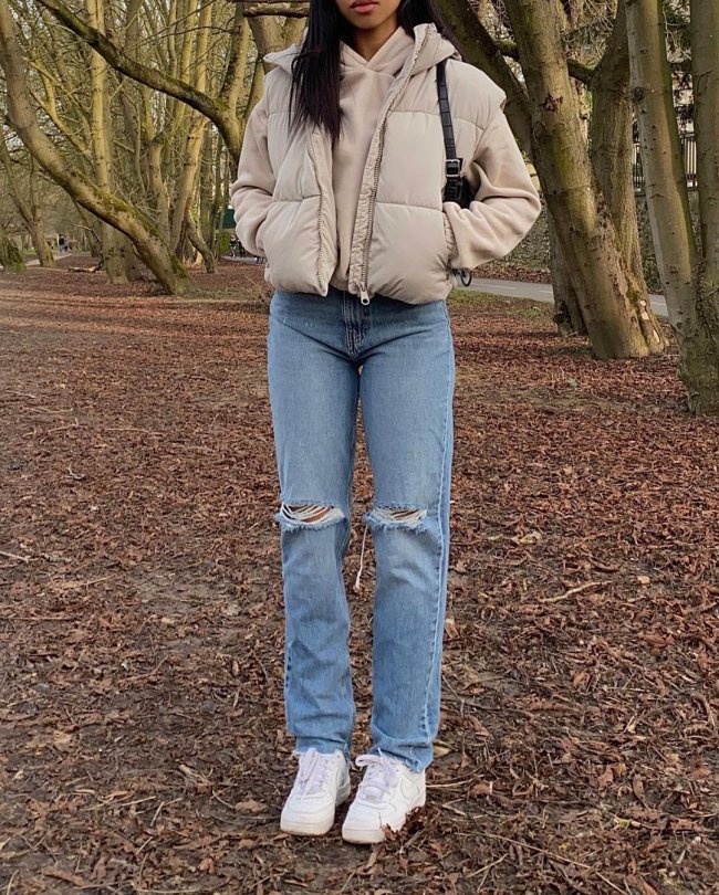 Foto do look de uma garota. Ela usa uma blusa de moletom bege, colete puffer bege, calça jeans e tênis branco. Ela está com as duas mãos no bolso do colete e não é possível ver o rosto dela na foto.