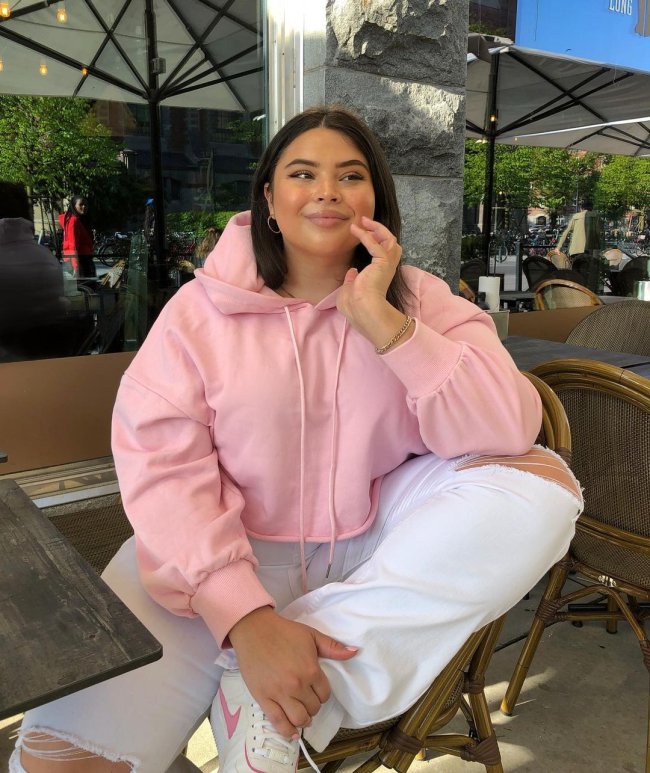 look-moletom-cortado Foto de uma mulher sentada em uma mesa de restaurante. Ela usa uma blusa de moletom com capuz rosa, calça jeans branca e tênis branco. Ela apoia a mão esquerda no queixo e a segura a perna esquerda com a mão direita, olha para o lado e sorri levemente.