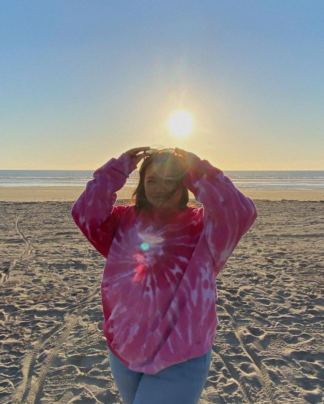 Foto de uma mulher na praia. Ela usa um moletom tie dye rosa, calça jeans e óculos de sol na cabeça. Ela segura o óculos de sol no topo da cabeça com as duas mãos, olha para a câmera e sorri levemente.