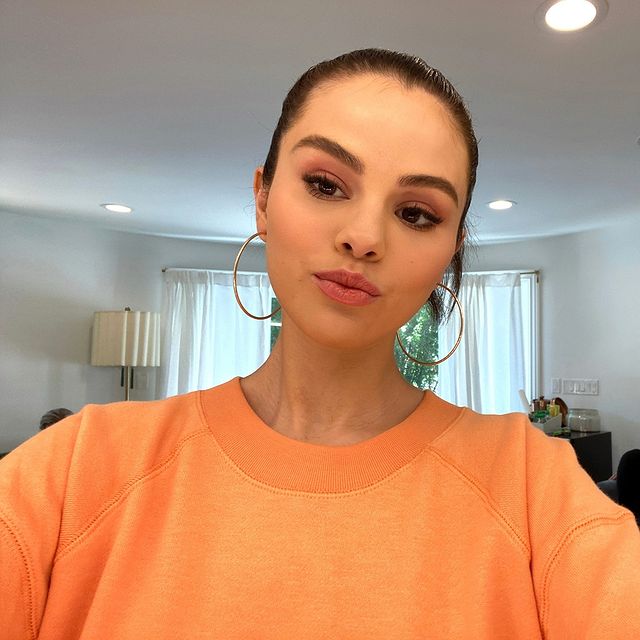 maquiagem-básica-selena-gomez Selfie da atriz e cantora Selena Gomez. Ela usa um moletom laranja, cabelo preso em um coque, brinco de argola dourado e maquiagem básica com olho esfumado e batom nude.
