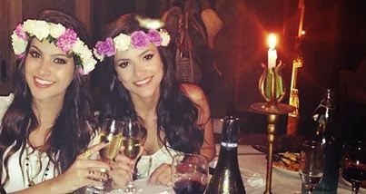 nah-manu-coroa-de-flores Foto da blogueira Nah Cardoso e cantora Manu Gavassi em um restaurante. Ambas usam uma blusa branca, coroa de flores e seguram uma taça de champanhe sorrindo.