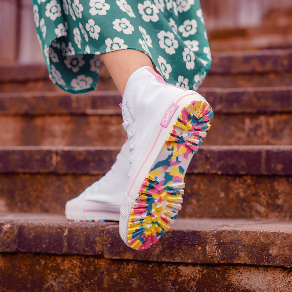 Foto do pé de uma menina usando tênis branco de cano alto da CAPRICHO Shoes com sola toda colorida. Ela está em um degrau de uma escada e dá para ver um pedaço do seu vestido longo verde com flores.