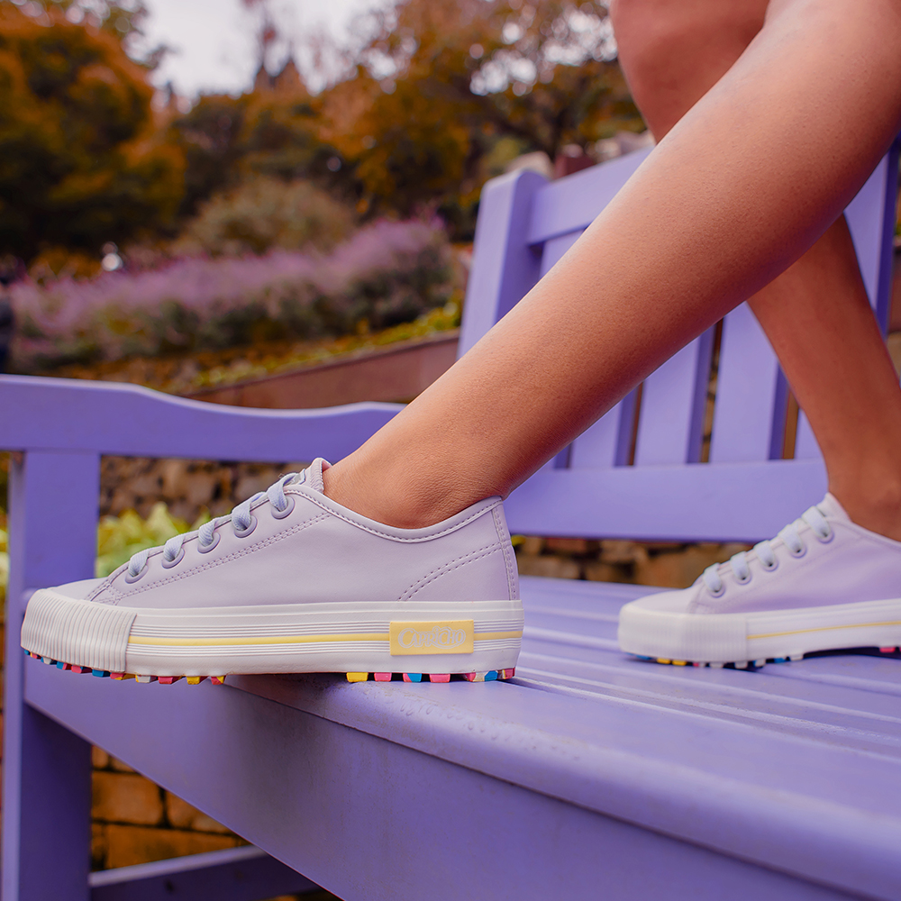 Garota usando tênis lilás com sola colorida da CAPRICHO Shoes. Na foto, dá para ver os pés dela, que estão apoiados em um banco lilás.