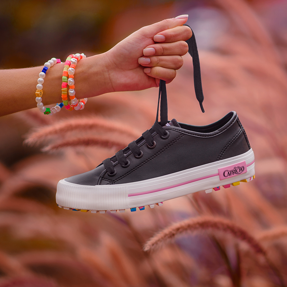 Garota segurando pelo cadarço o tênis preto com branco e sola colorida da CAPRICHO Shoes. Na foto, dá para ver apenas uma de suas mãos segurando o sapato.