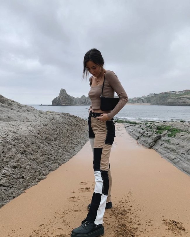 Jovem posando na praia com blusa marrom e calça com patchwork com detalhes em preto e marrom, ela olha para baixo e uma de suas mãos segura o cós da calça.