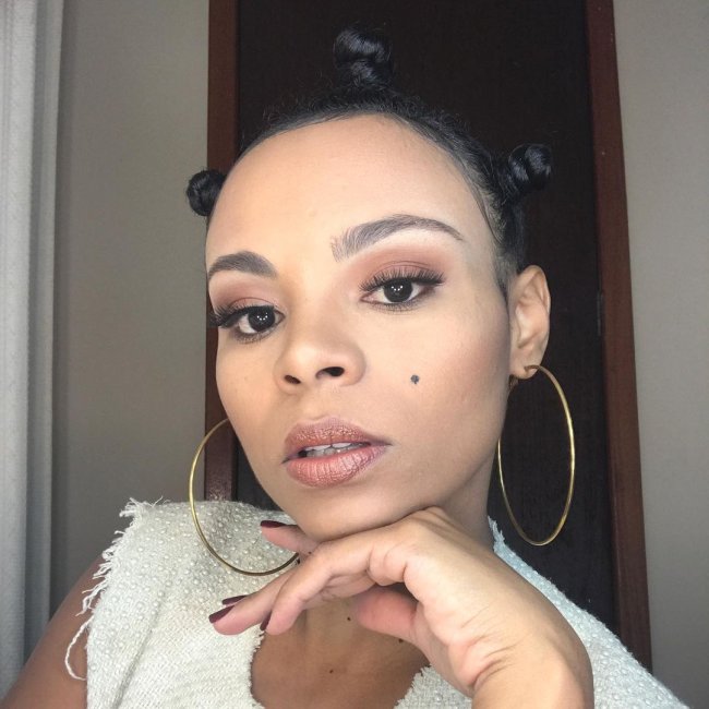 penteado cabelo cacheado Jovem posando para selfie com expressão séria, ela apoia sua mão direita no queixo e usa brincos de argola.