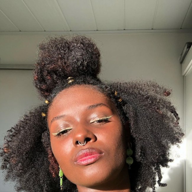 penteado coque meio preso cabelo cacheado curto Jovem posando para selfie, só conseguimos ver seu rosto. Ela tem uma expressão séria e usa coque no topo da cabeça.