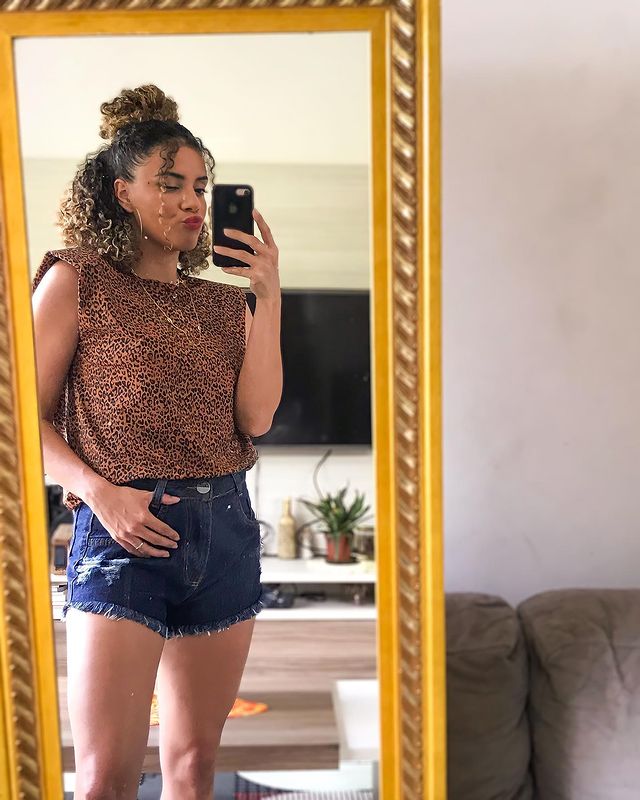 penteado meio preso cacheado Jovem tirando foto no espelho com short jeans e camiseta. Sua expressão é sorridente.