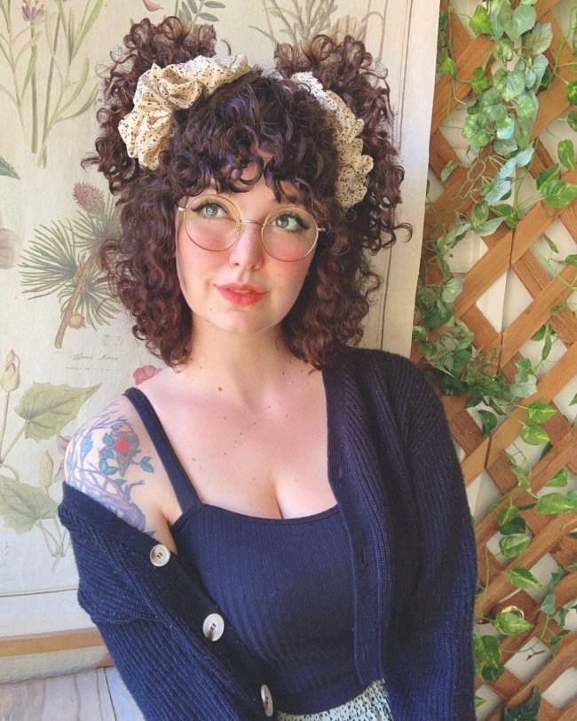 scrunchies em cabelo cacheado Jovem com expressão sorridente usando óculos, regata azul e cardigan.