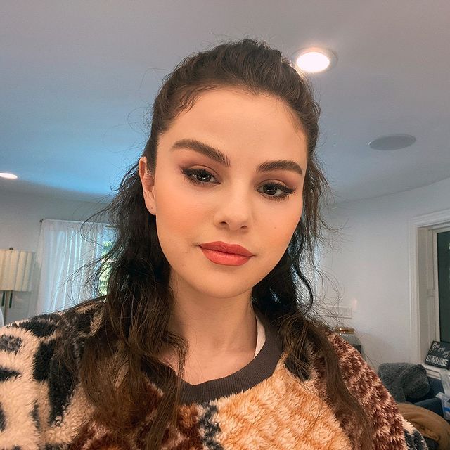 Selfie da cantora e atriz Selena Gomez. Ela usa um casaco de pelinhos marrom, cabelo meio preso e maquiagem com olho esfumado e batom nude. Ela olha para a câmera e sorri levemente.