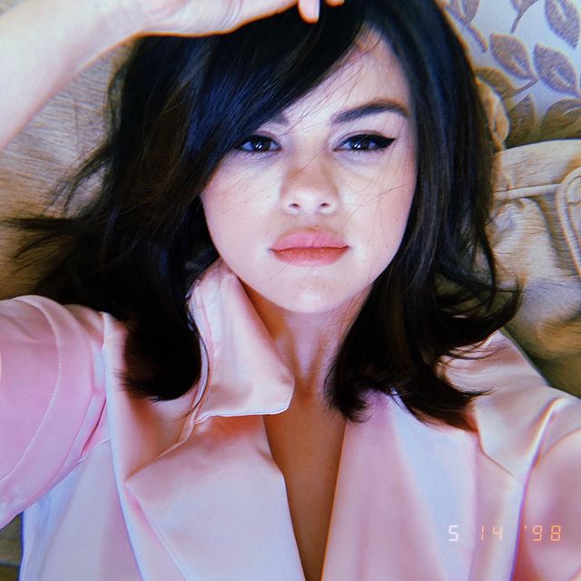 Selfie da cantora e atriz Selena Gomez. Ela usa uma camisa rosa, cabelo solto, maquiagem natural com delineado gatinho preto. Ela olha para a câmera, não sorri e segura o cabelo com a mão direita.