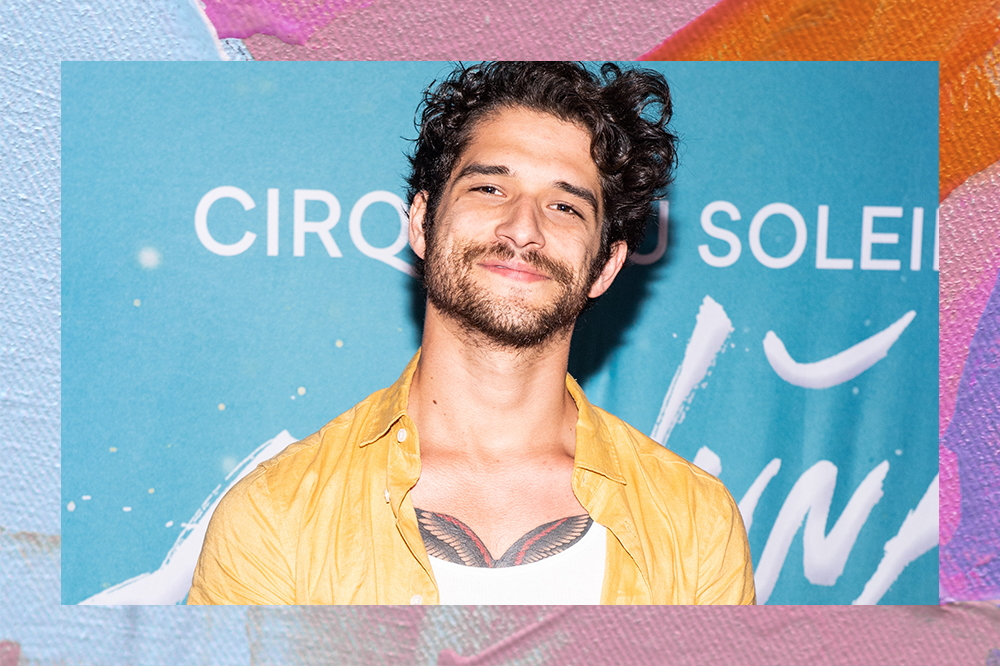 Tyler Posey com uma camisa amarela aberta, um leve sorriso. Ao fundo um painel azul com lestras brancas