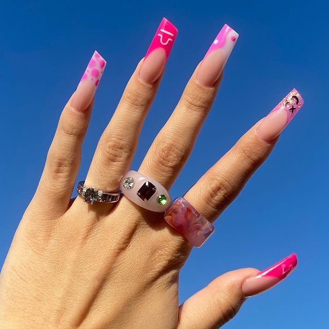 Foto de uma mão com o céu como fundo. A mão está com unhas decoradas em formato de batom e três anéis divertidos.