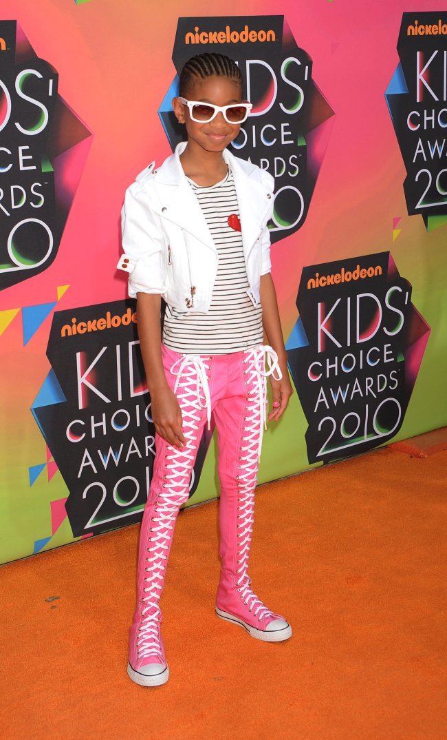 Foto da atriz e cantora Willow Smith no Kids Choice Awards 2010. Ela usa um camiseta listrada, casaco branco, calça tênis rosa All Star e óculos de sol branco. Ela olha para a câmera e sorri.