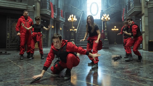 Cena da quinta temporada de La Casa de Papel; os personagens estão na área central do lobby do Banco da Espanha usando os macacões vermelhos com camisetas pretas e fazem gestos de apreensão e cuidado abaixando as armas; Palermo aparece agachado encarando algo na frente do grupo colocando sua arma no chão