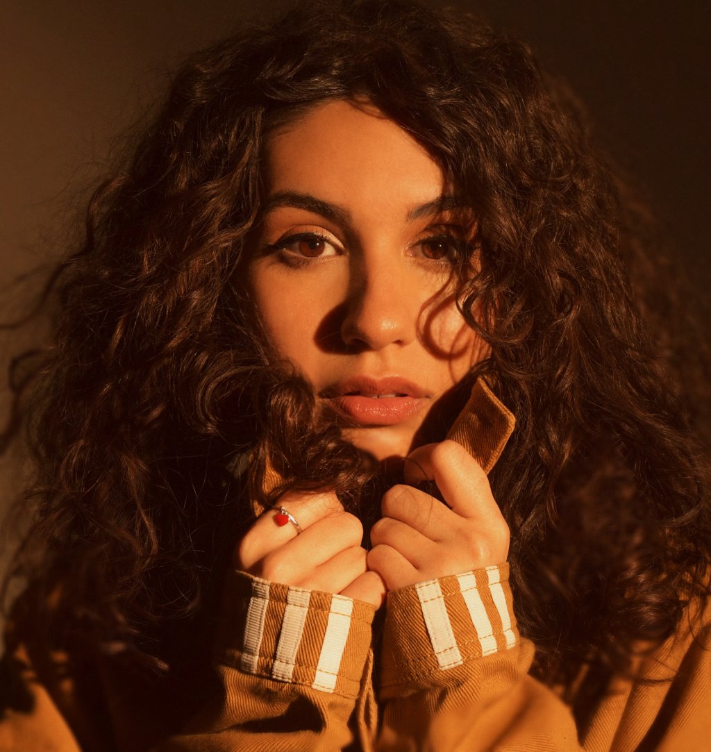 Alessia Cara em foto de divulgação com tons mais quentes e amarelados; ela está segurando a gola do casaco com as duas mãos e escondendo o pescoço e parte do rosto; a expressão da cantora é séria