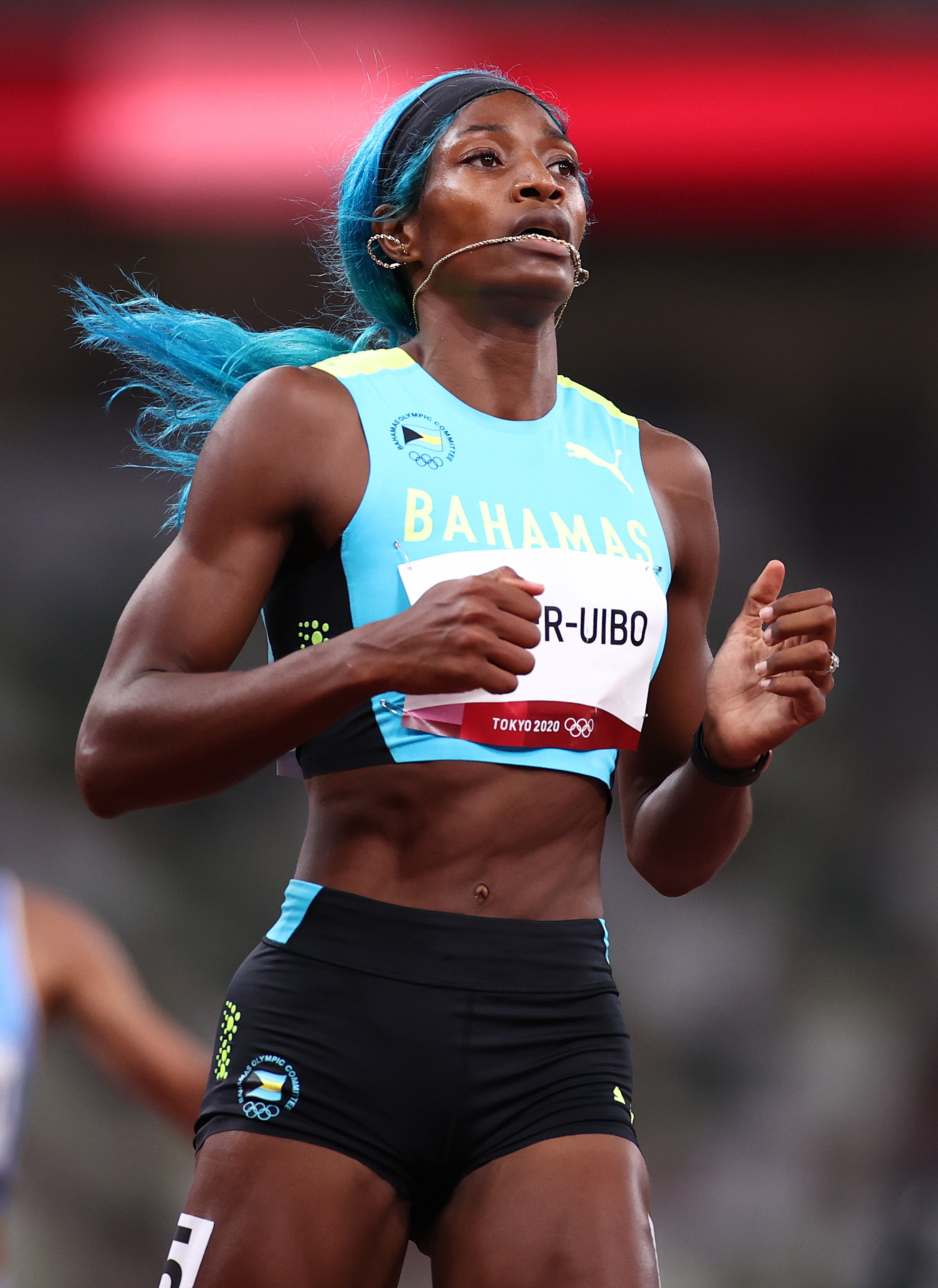 Shaunae Miller-Uibo Shaunae Miller-Uibo, das Bahamas, na pista de atletismo das Olimpíadas de Tóquio 2020. Ela está correndo, usando uma lace azul e com um colar na boca.