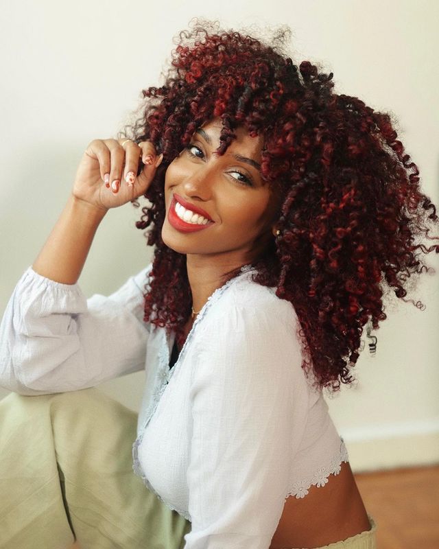 cabelo-cacheado-vermelho Foto de uma mulher. Ela usa uma blusa de manga bufante branca, maquiagem natural com batom vermelho, cabelo cacheado solto em um tom de vermelho. Ela está sentada em um sofá, olha para a câmera e sorri.