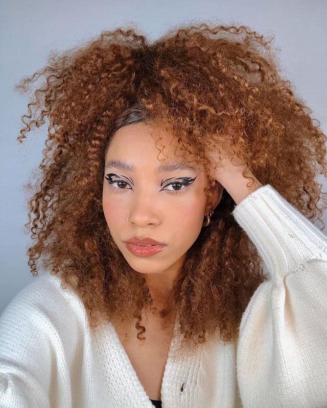 cabelo-ruivo-acobreado-crespo Selfie de uma mulher. Ela usa um cardigan branco, maquiagem com delineado gráfico preto e cabelo crespo meio preso em um tom de ruivo acobreado. Ela está com a mão esquerda no cabelo, olha para a foto e não sorri.